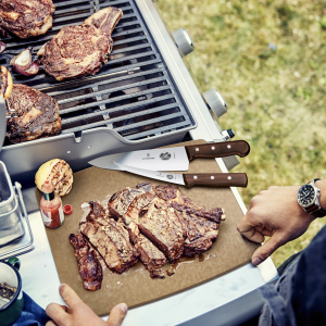 Top down shot of a grill with meat on it and two knives on a cutting board (one being a victorinox boning knife)